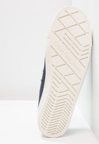 Calvin Klein Jeans sneaker sula, med en vit gummisula som har ett texturerat greppmönster och en m örk tygöverdel med metallaccenter.