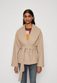 Μπεζ, υπερμεγέθης παλτό περιτυλίγματος με φαρδύ γιακά και ζώνη, φτιαγμένο από μαλακό υφασμάτινο υλικό. Φορεμένο �πάνω από ένα πουλόβερ με πολύχρωμο σχέδιο και τζιν.