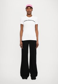 Balts pieguļošs T-krekls ar melnā krāsā izšūtu uzrakstu "Versace Jeans Couture", apvienots ar brīviem melniem biksēm un rozā cepuri.