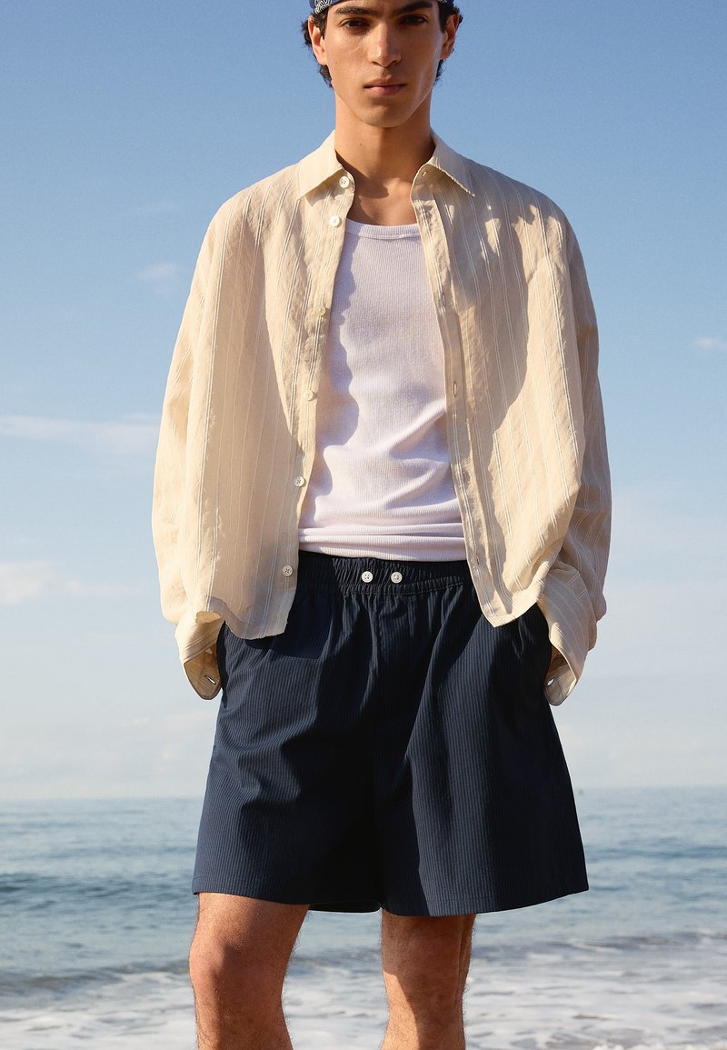 Jeune homme debout au bord de la mer, portant une chemise beige clair ouverte, un débardeur blanc et un short bleu marine foncé, les mains dans les poches.