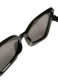 Ochelari de soare negri cu un design geometric. Rame groase, lucioase și lentile întunecate. Unghiuri notabile la sprânceană și tâmple proeminente.