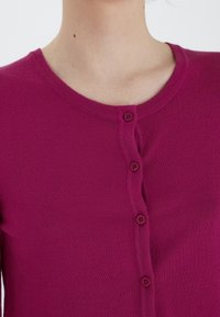 Fuksjowy kardigan z okrągłym dekoltem, zapinany na guziki z przodu, wykonany z miękkiej, teksturowanej dzianiny.