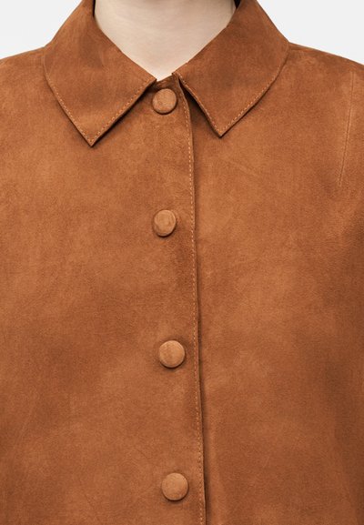 Καφέ πουκάμισο σουέντ με αιχμηρό γιακά και τέσσερα κουμπιά καλυμμένα με ύφασμα στο μπροστινό μέρος, που φοριέται από ένα άτομο μπροστά σε απλό φόντο.