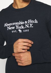 Maglione lavorato a maglia blu navy con scollo rotondo, caratterizzato da testo ricamato bianco e logo di alce. Dettagli a coste sui polsini e sull'orlo.