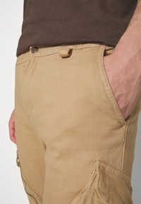 Gros plan sur une personne portant un pantalon cargo beige avec la main dans la poche latérale et une chemise marron foncé rentrée.