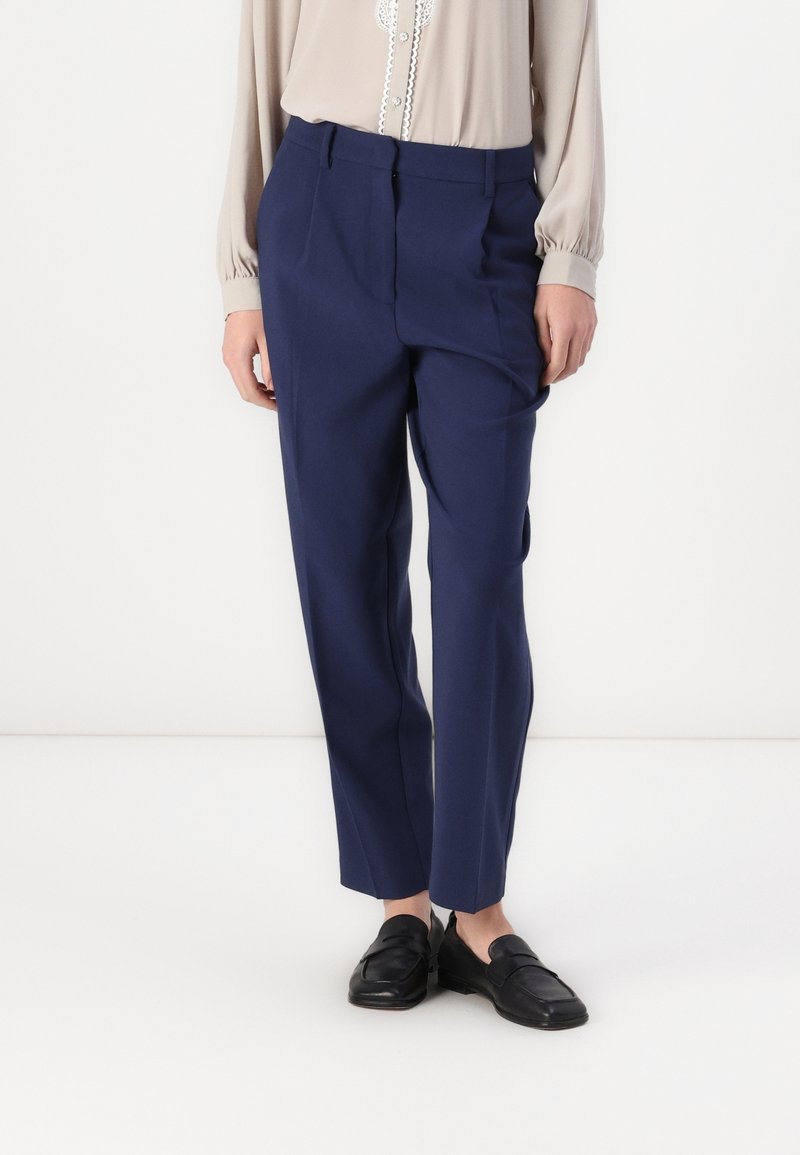 Personne portant un pantalon bleu marine sur mesure, un chemisier beige à manches longues avec bordure en dentelle, et des mocassins en cuir noir sur fond blanc.