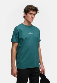 Teal bomull t-skjorte med korte ermer, rund hals, og hvit trykt logo på brystet; sammen med svarte bukser.