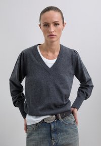 Femme aux cheveux châtain clair attachés, portant un pull à col V gris foncé par-dessus une chemise blanche et un jean bleu avec une ceinture noire, devant un fond uni.