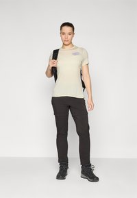 Light beige short-sleeve T-shirt with purple text, paired with dark, tapered pants and black hiking boots, worn with a black backpack.