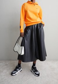 Sudadera naranja, falda de cuero sintético negra, bolso blanco y negro, y zapatillas negras/grises. Detalles destacados como el cordón y materiales texturizados.