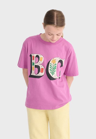 Ung person iført en lilla T-shirt med store bogstaver "B" og "C" dekoreret med blomster, som kigger ned med hænderne bag ryggen.