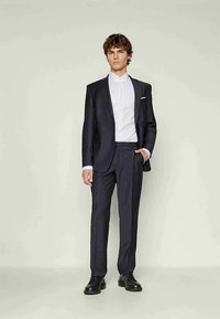 Fato escuro bem ajustado com um design estruturado, lapelas elegantes, calças a condizer, camisa branca de colarinho e sapatos pretos. Textura de tecido suave.
