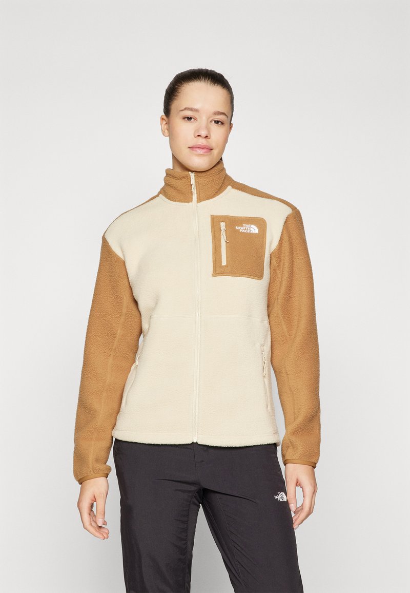 Fleece striukė su kreminiu korpusu ir rudomis rankovėmis. Yra užtrauktuku užsegama krūtinės kišenė, aukšta apykaklė ir prekės ženklo logotipas. Minkšta tekstūra.