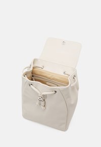 Beige bucket-väska med dragsko och klaffstängning, öppen för att visa beige tygfoder och ett innerfack med dragkedja.