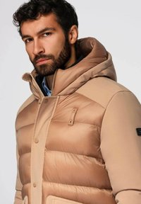 Beige pufferjack met een capuchon, voorzien van een geluidsontwerp, metalen drukknoopsluitingen en vo pockets. Glad textuur en minimalistische accenten.