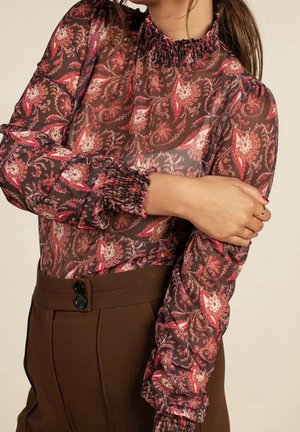 Kobieta w przezroczystej, długiej, kwiecistej bluzce w odcieniach czerwieni i brązu, w połączeniu z wysokim stanem ciemnobrązowych spodni z dwoma guzikami.