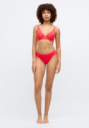 Frau mit lockigem Haar, die roten Spitzen-BH und passende Unterwäsche trägt, steht barfuß vor einem schlichten weißen Hintergrund.