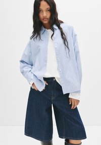 Lyseblå stribet skjorte med knaplukning i en oversized pasform, parret med mørk denim culottebukser. Har en hvid langærmet undertrøje og knæhøje støvler.