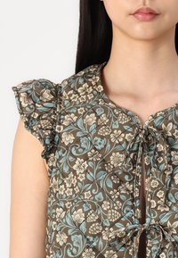 Braune Bluse mit floralem Muster und Puffärmeln, ausgestattet mit einem Bindeband, filigranen blau-weißen Mustern und einer gesteppten Textur.