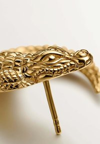 Hebilla para el cabello en forma de cocodrilo dorado que presenta escamas y rasgos faciales detallados, con un prong resistente para asegurar el cabello. Acabado metálico suave.