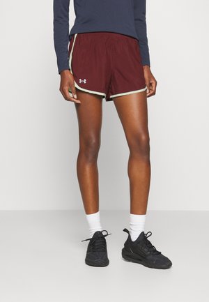 Persoon staat binnen en draagt bordeauxrode sportshorts met witte bies, donkerblauwe lange mouwen shirt, witte sokken en zwarte hardloopschoenen.
