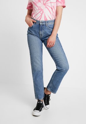 Persoon die hoge blauwe jeans, een roze-wit tie-dye t-shirt en zwarte hoge sneakers draagt, staat met één hand in de zak.
