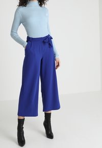 Un col roulé côtelé bleu clair associé à un pantalon large royal bleu taille haute avec un nœud, porté avec des bottes à talons noires.
