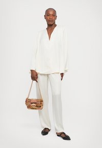 Mujer calva vestida con una blusa blanca suelta y pantalones, sosteniendo un bolso tejido multicolor, de pie contra un fondo blanco.