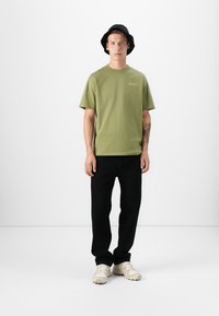 Zaļš kokvilnas T-krekls ar apaļu kaklu un dzelteno logotipu, apvienots ar melniem biksēm un bežām kurpēm, valkāts ar melnu cepuri uz vāciņa.