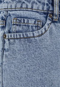 Le tissu en denim bleu clair présente une texture subtile avec un tissage diagonal. Le détail de la poche comprend des coutures jaunes et un accent de rivet en métal.