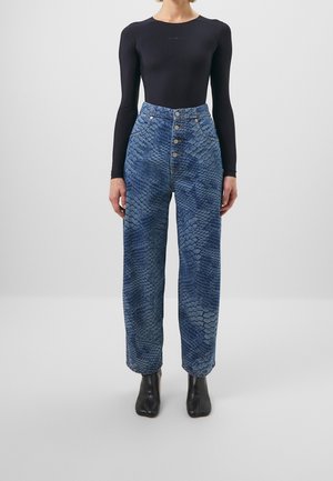 Persona que lleva vaqueros azules de tiro alto con estampado de serpiente, camiseta negra de manga larga y botines negros, de pie frente a un fondo neutro.