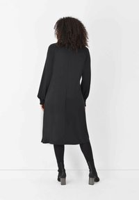 Svart midiklänning med långa ballongärmar, slät textur och rak skärning. Har en söm bak och bärs med ankelstövlar.