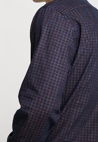 Camicia da uomo a maniche lunghe in blu scuro e bordeaux a quadri, con un design aderente e tela strutturata. Dettaglio di cuciture visibili.