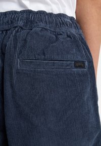 Pantaloni corti di velluto a coste blu scuro con cintura elastica e una tasca posteriore con un piccolo etichetta nera. Strisce verticali testurizzate sono visibili.