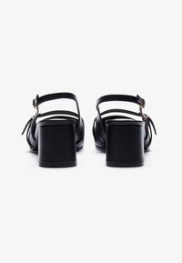 Sandali in pelle nera con tacco a blocco. Presentano una cinghia alla caviglia con fibbia e sottili cinghie attraversate sul piede, texture liscia, design semplice.