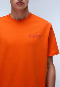 Orange bomulds t-shirt med rund halsudskæring. Har et broderet logo i lilla på brystet. Korte ærmer og glat tekstur.