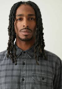 Camicia a scacchi grigia con bottone, caratterizzata da una tasca frontale, colletto e tessuto testurizzato. Il design include linee orizzontali e verticali.