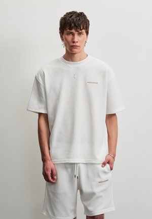 Young man with short hair wearing white ribbed oversized t-shirt and matching shorts, standing against plain light background.