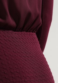 Vêtement bordeaux avec une ceinture texturée ornée de motifs en relief ; tissu doux et fluide au-dessus de la taille, créant un effet superposé.