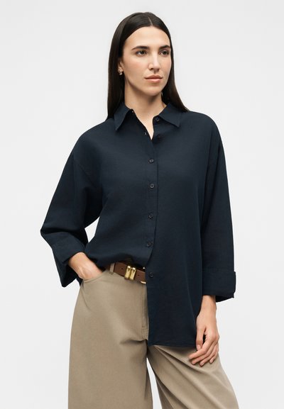 Γυναίκα με μακριά σκούρα μαλλιά, φορώντας ένα φαρδύ σκουρομπλε πουκάμισο με κουμπιά και μπεζ παντελόνια με καφέ ζώνη, ποζάρει με το ένα χέρι στην τσέπη.