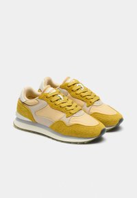 Zapatillas deportivas que combinan una parte superior de ante amarillo y malla, detalles en beige, texturas variadas y una media suela blanca con suela exterior gris.