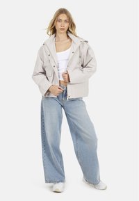 Veste imperméable beige clair avec capuche, fermeture éclair et accents en boutons, associée à un jean en denim clair ample et des chaussures blanches.