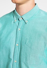 Türkisfarbenes Hemd aus leichtem Stoff mit Knopfleiste, Kragen, Brusttasche und hellen Knöpfen. Glatte Textur mit feiner Webstruktur.