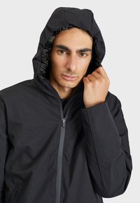 Čierna kapucňová bunda s hladkou textúrou, so zapínaním na zips vpredu a naťahovacou kapucňou. Diskrétne detaily okolo goliera a manžiet.