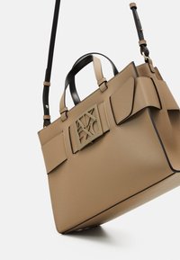 Béžová strukturovaná kabelka s černými prvky a horními rukojeťmi. Má výrazné logo a přední kapsy s klopou, vyrobená z hladké kůže.