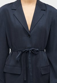 Κοντινή εικόνα ενός ατόμου που φοράει μπλέ σακάκι με ένα μόνο μαύρο κουμπί και μια υφασμάτινη ζώνη δεμένη στη μέση.