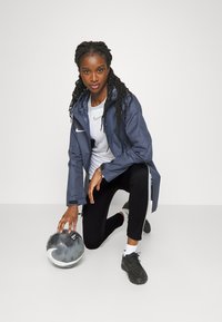 Marine regenjack met capuchon en ritszak, wit Nike-logo, gelaagd over een wit shirt, zwarte leggings en zwarte sport schoenen, knielend bij een voetbal.