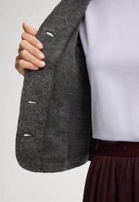 Giacca di lana grigia tenue aperta per rivelare un morbido rivestimento interno bianco. Dettagli notevoli includono asole sui bordi e una vestibilità comoda.
