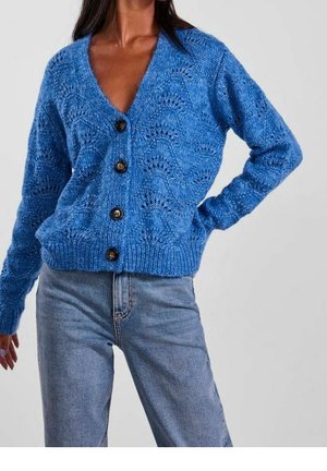 Femme portant un cardigan bleu tricoté avec des boutons noirs et un jean bleu clair, posant avec une main sur la hanche et un bras détendu.