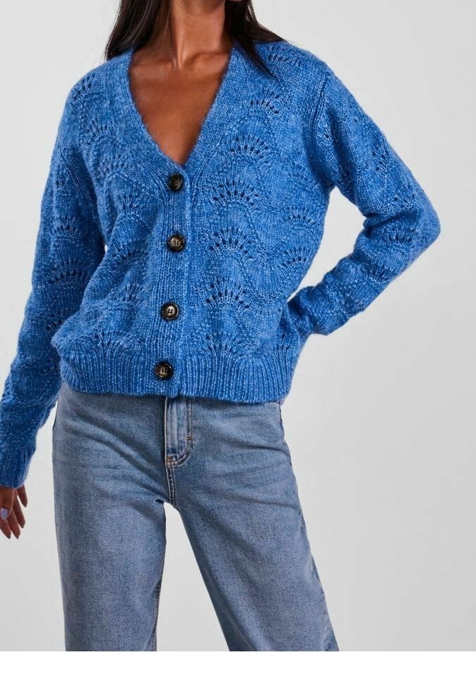 Femme portant un cardigan bleu tricoté avec des boutons noirs et un jean bleu clair, posant avec une main sur la hanche et un bras détendu.